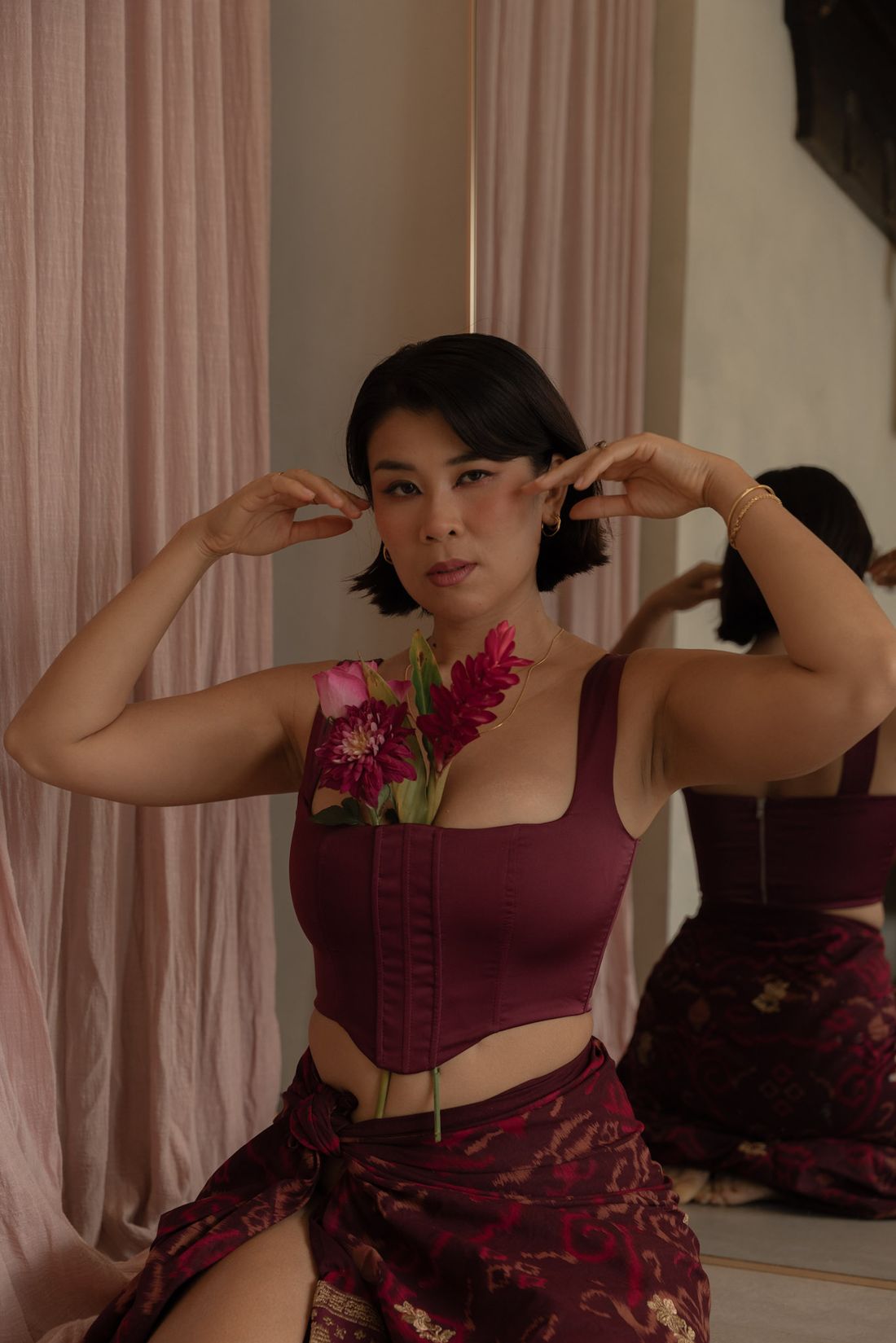 Nadia in a deep burgundy corset with flowers tucked at her chest, hands framing her face