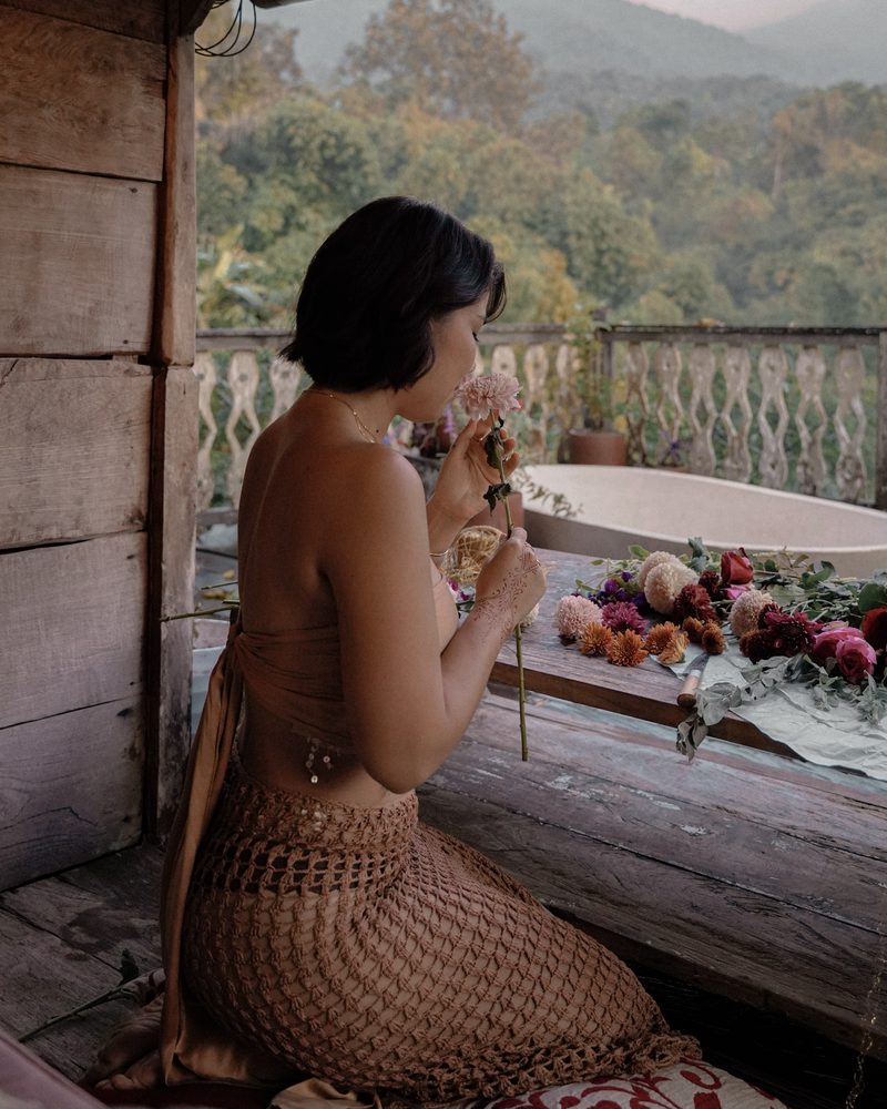 A woman kneeling in a mountain cabin, smelling a flower beside a table of blooms