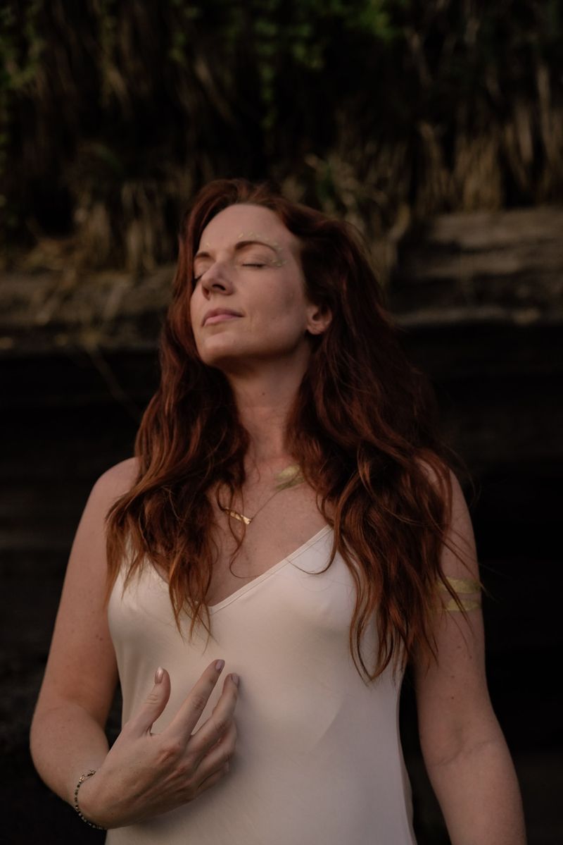 A woman with long red hair, eyes closed, one hand resting on her chest